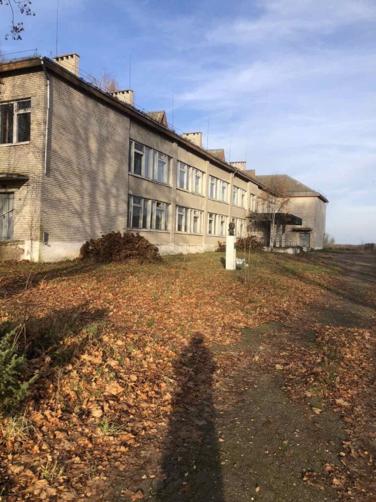 Под голубым небом на покрытом опавшими осенними листьями травянистом склоне стоит двухэтажное кирпичное здание с множеством окон. На переднем плане, на тропинке, видна тень человека.