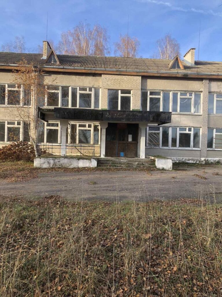 Заброшенное двухэтажное здание с разбитыми окнами и обветшалым фасадом, окруженное заросшей травой и осенними деревьями под голубым небом.